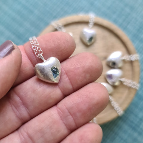 Silver heart-shaped pendant with a blue stone held between fingers against a teal background.