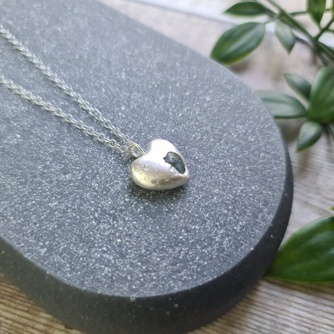 Silver heart-shaped pendant necklace on a dark gray stand with green leaves in the background