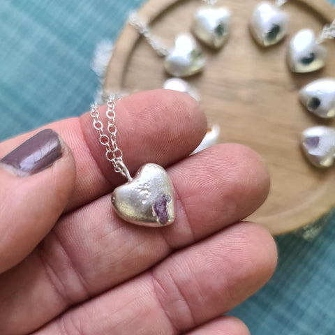 Heart-shaped pearl pendant on a chain held by a hand with a blurred background of more pendants.