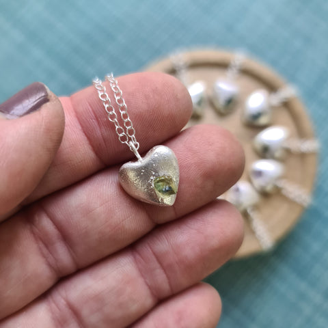 Silver heart-shaped pendant with a green stone held between fingers against a blue background.