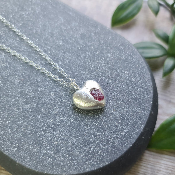 Silver heart-shaped pendant with a red ruby gemstone on a dark surface with green leaves in the background