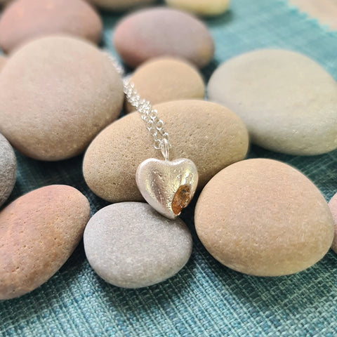 Heart-shaped silver pendant with a small orange gemstone on a necklace, lying on pebbles.
